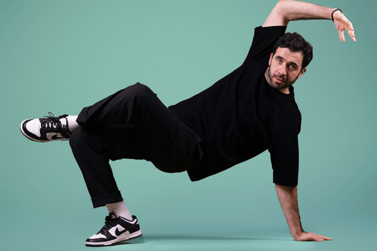 Man in zwarte kleiding staat op 1 been en 1 arm met groenige achtergrond