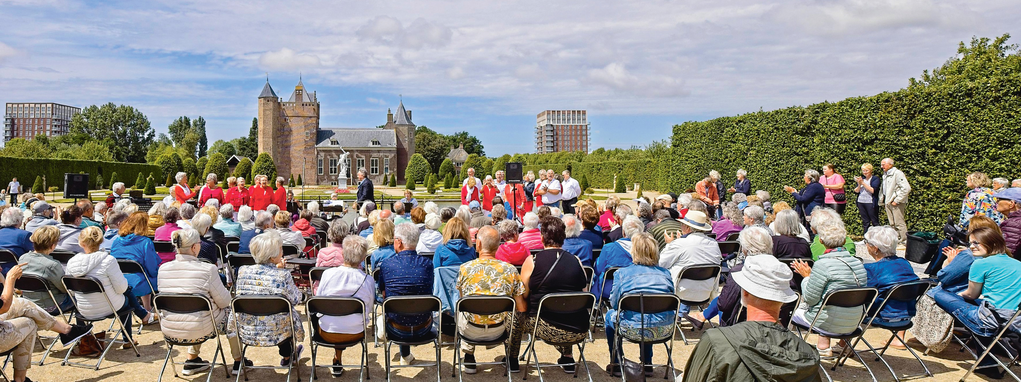 Een grote groep mensen zit buiten op stoelen voor Slot Assumburg te luisteren naar een zangkoor.