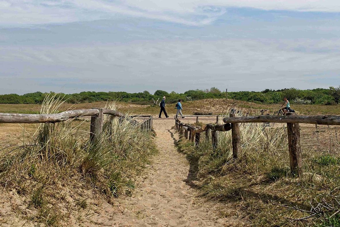 Twee wandelaars op een zandpad in de duinen met op de voorgrond een houten hek.